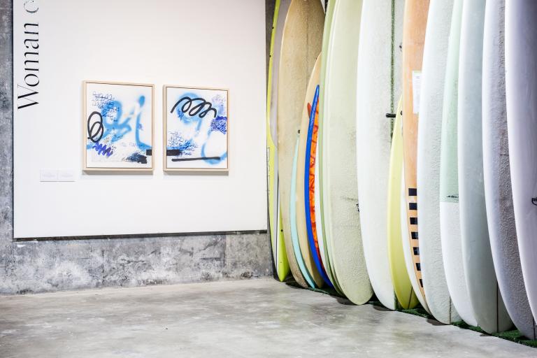 Image of gallery with surfboards lined along the right hand side and two bllue ink based artworks hung on the wall.