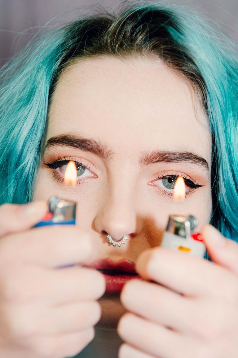 A lady with blue hair holding two lighters in front of her eye